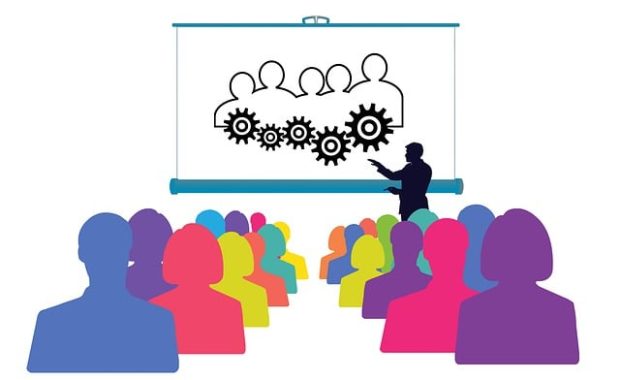 human silhouettes in diverse colours arranged in a training setting with a presentation in front and a presenter by the side.